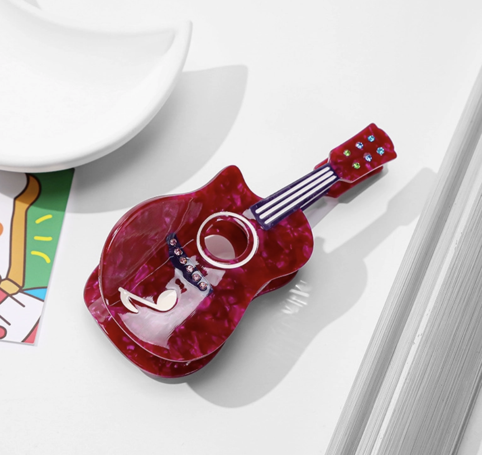 A glossy red guitar-shaped hair clip with a marbled pattern, featuring a circular cutout in the center and small colorful embellishments on the neck, photographed on a white surface with partial props around it.