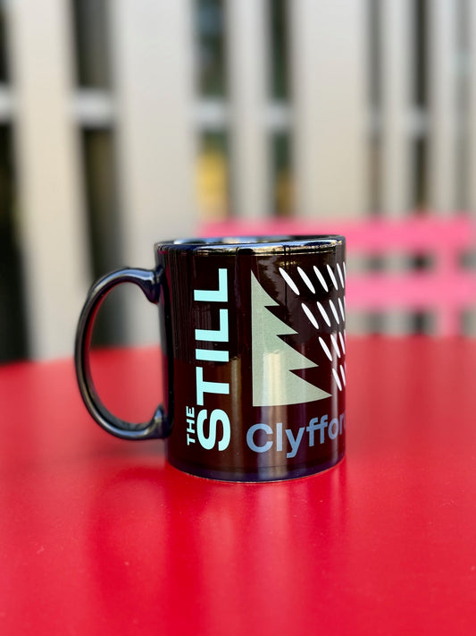 Black mug with 'The Still Clyfford' logo on a red surface