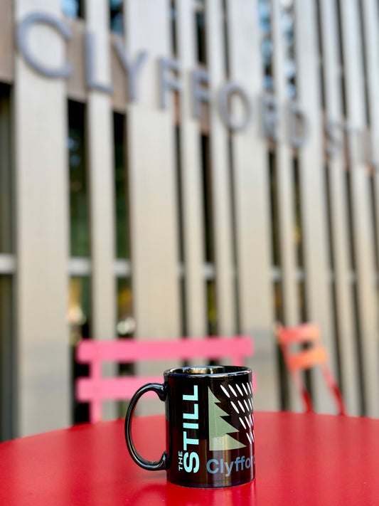 Black mug with 'The Still' branding on a red table in front of a blurred Clyffe sign.