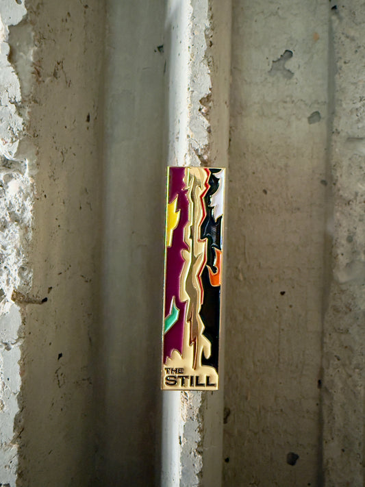 Colorful lapel pin  with 'The Still' branding against a concrete wall.