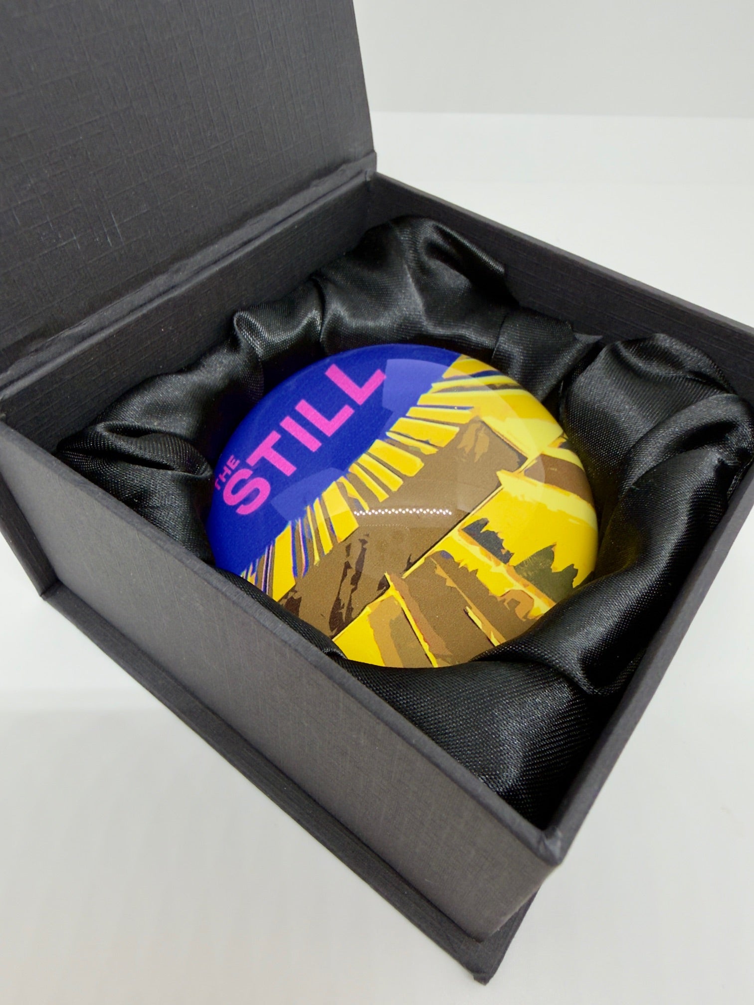 Colorful round paperweight with 'THE STILL' logo in a black box