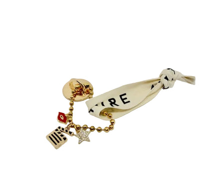A gold-tone charm keychain featuring a round pin-back clasp with multiple dangling charms, including a small red lips, a striped rectangular charm, a star charm, and a cream ribbon tag printed with black text, shown on a light gray background.