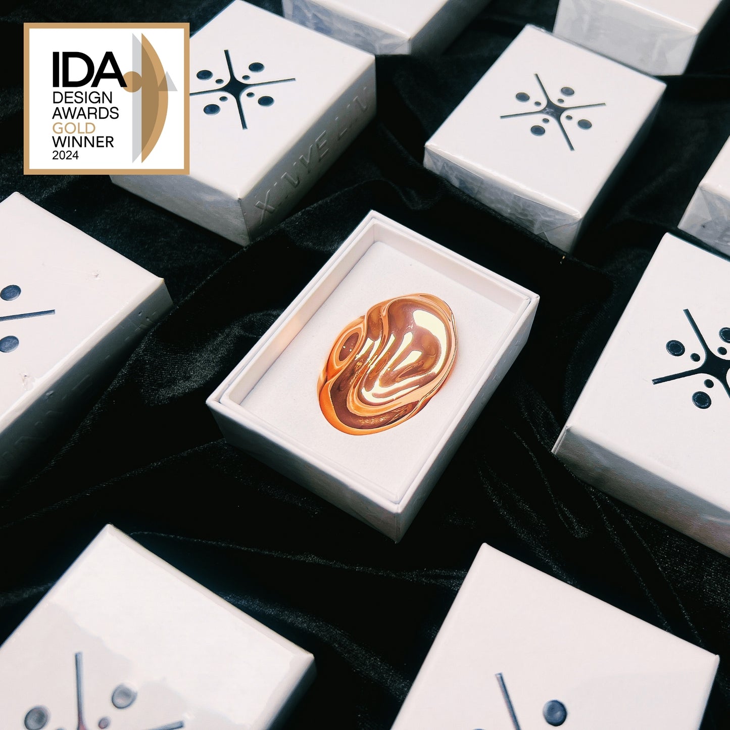 Gold solid perfume in a box surrounded by white boxes with black designs, on a dark fabric background.