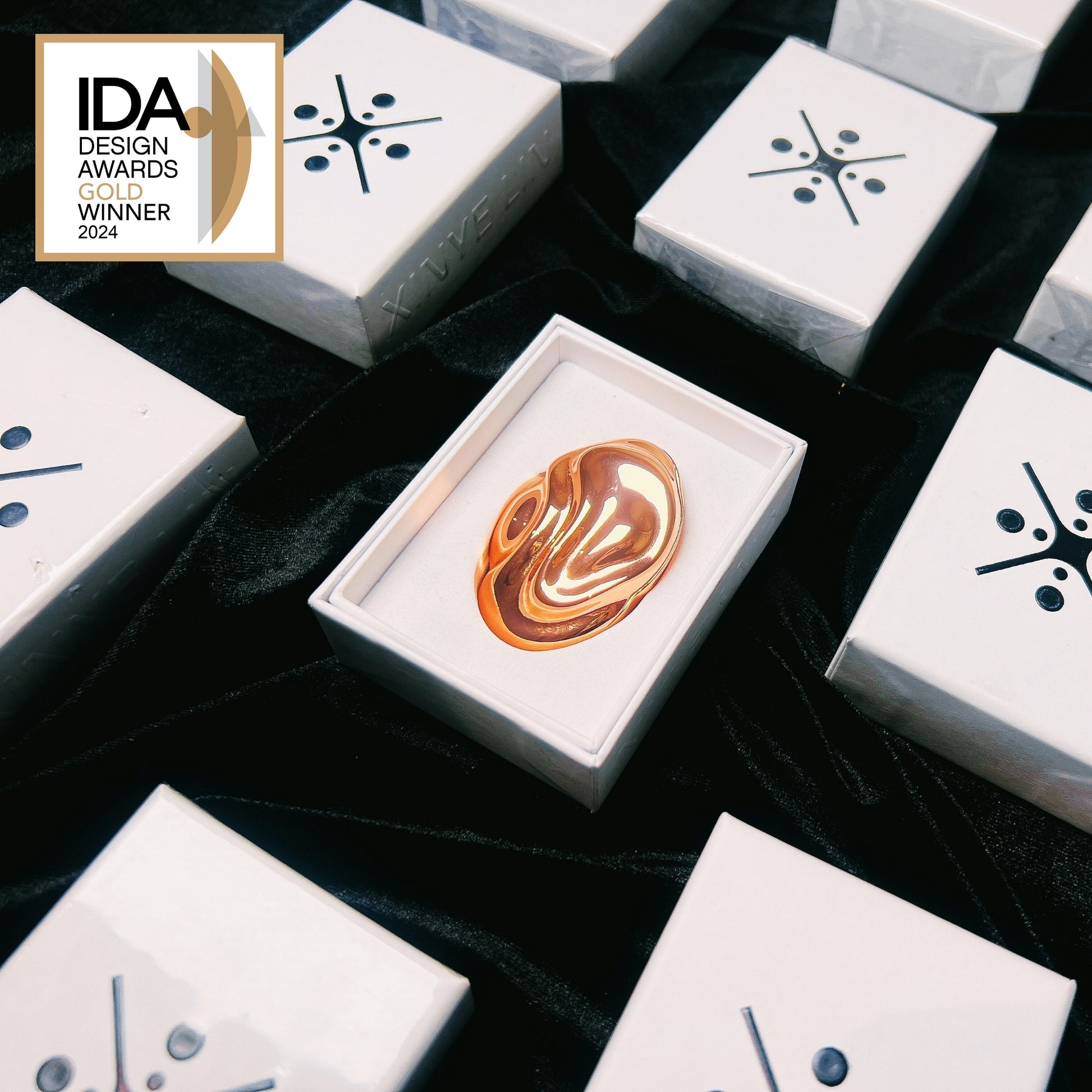 Gold solid perfume in a box surrounded by white boxes with black designs, on a dark fabric background.