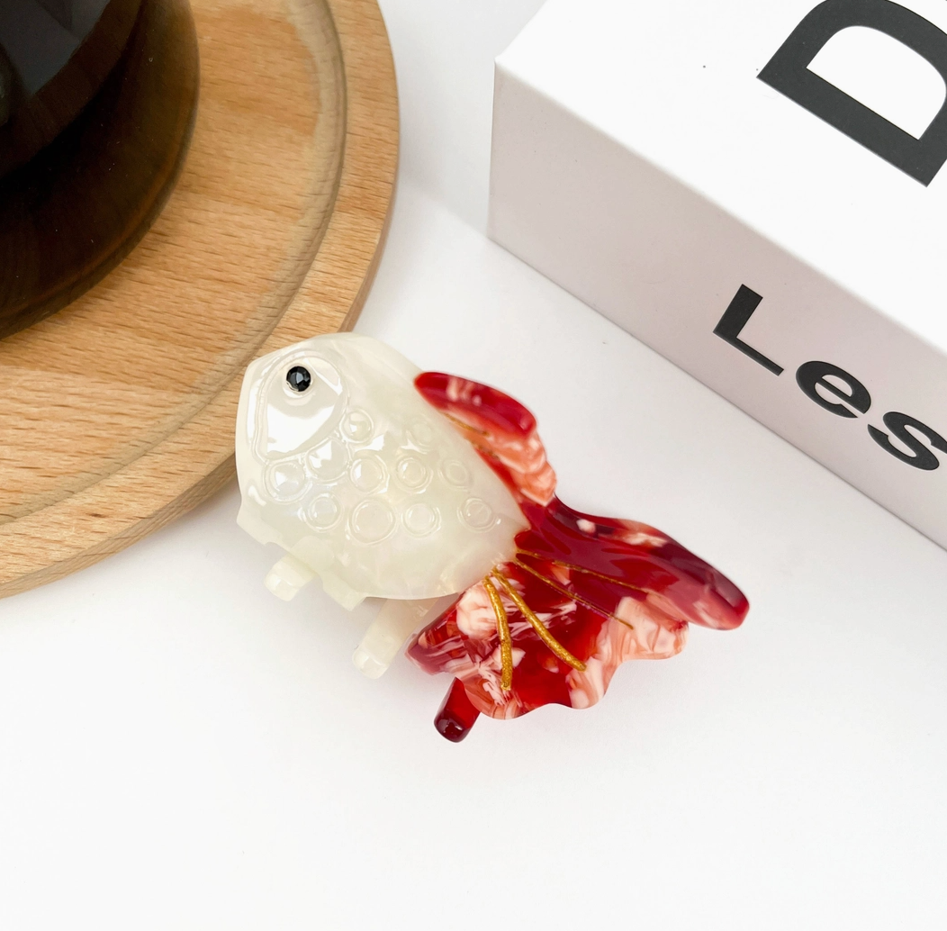 Decorative goldfish hair clip with a white body and red tail on a white surface.