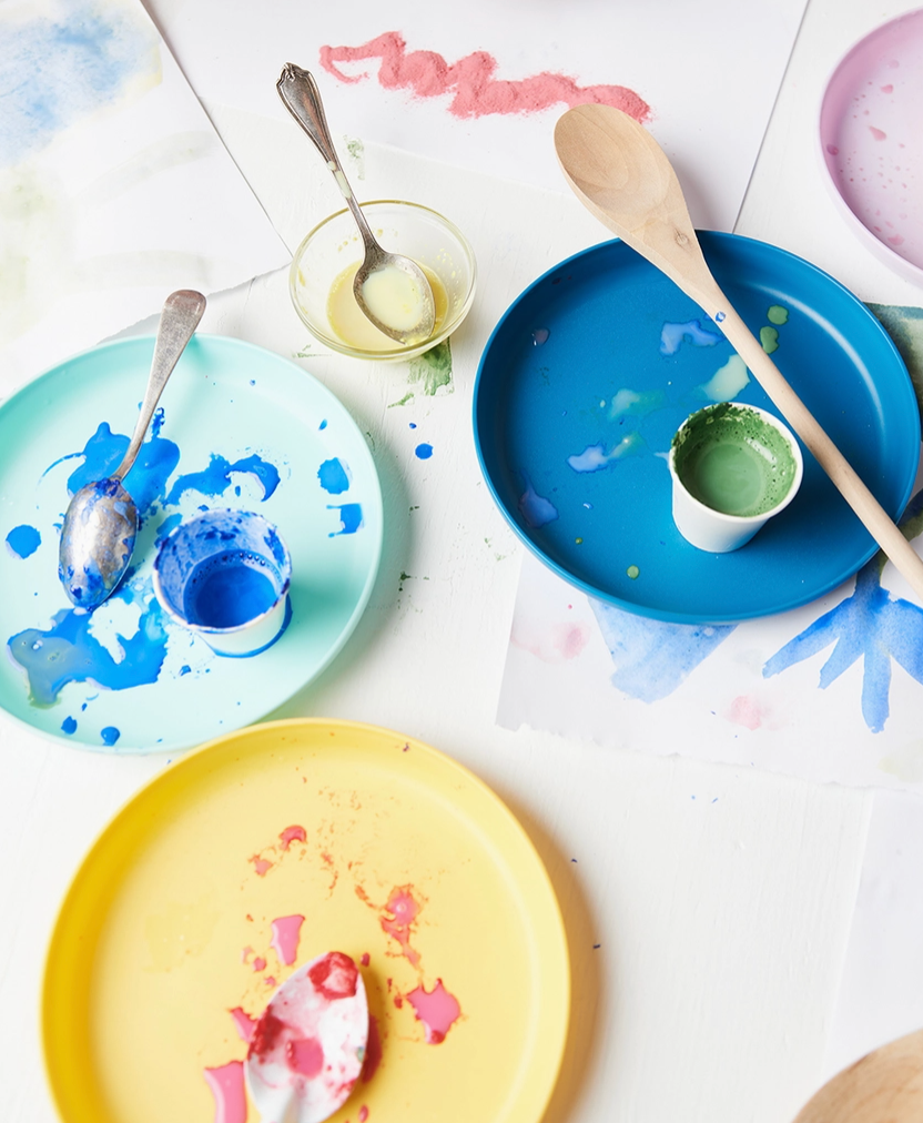 Colorful plates with paint splashes and utensils on a white surface.