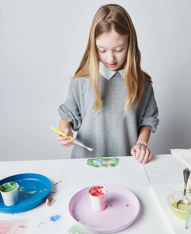 Young girl painting a small cupcake-shaped object with a brush on a white table.