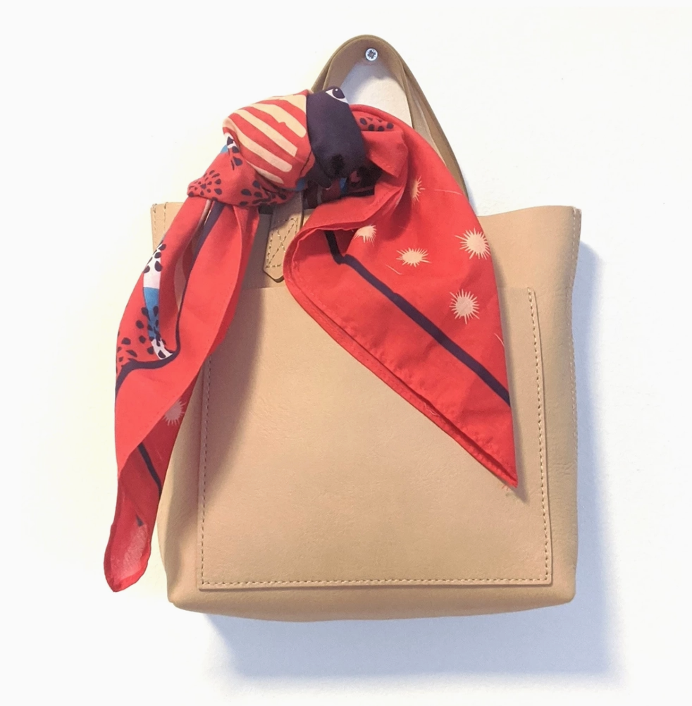 Beige handbag with a red patterned scarf tied around it on a white background