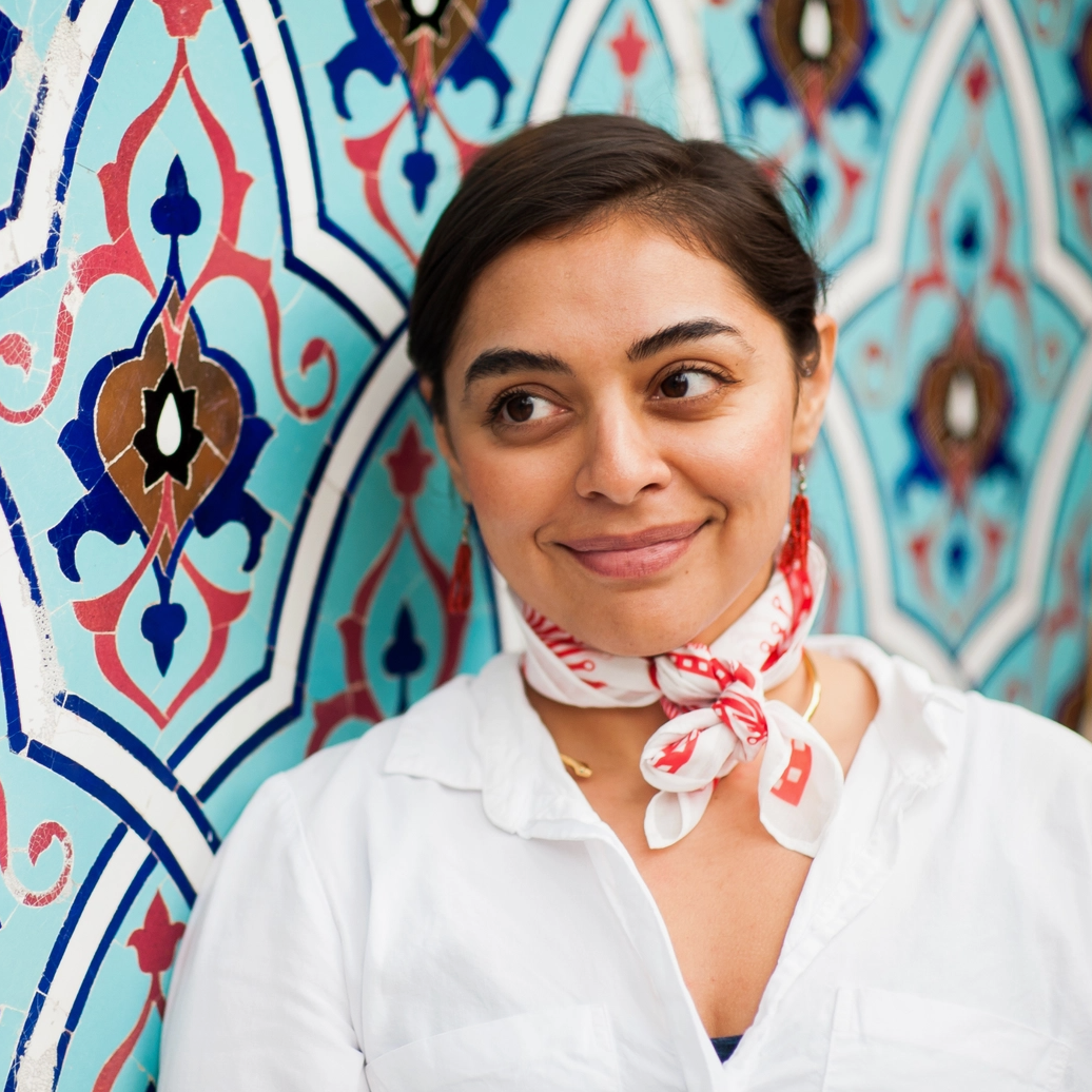 Woman wearing a white shirt with a red and white patterned scarf against a decorative wall.