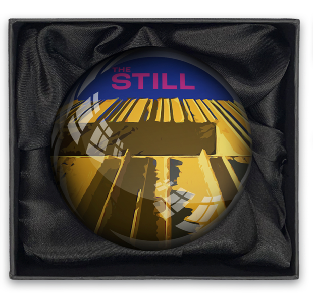 Round glass paperweight with 'The Still' branding inside a black box