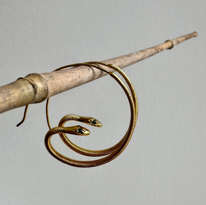 Gold snake-shaped earrings on a wooden stick against a gray background