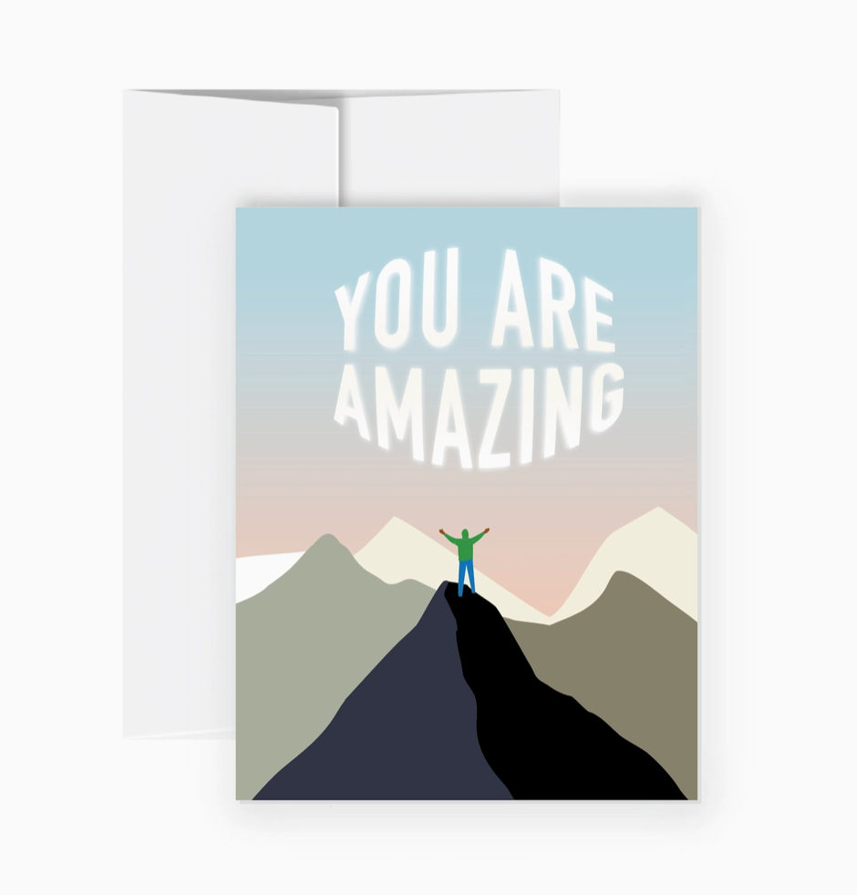 A greeting card with a white envelope behind it, featuring the text 'YOU ARE AMAZING' above a minimalist mountain landscape with a small person standing on a peak.