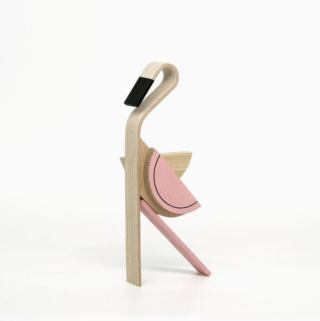 A modern wooden bird sculpture or decorative object with layered plywood construction, featuring a natural wood body, a pink wing, and a black accent on the beak, set against a plain white background.