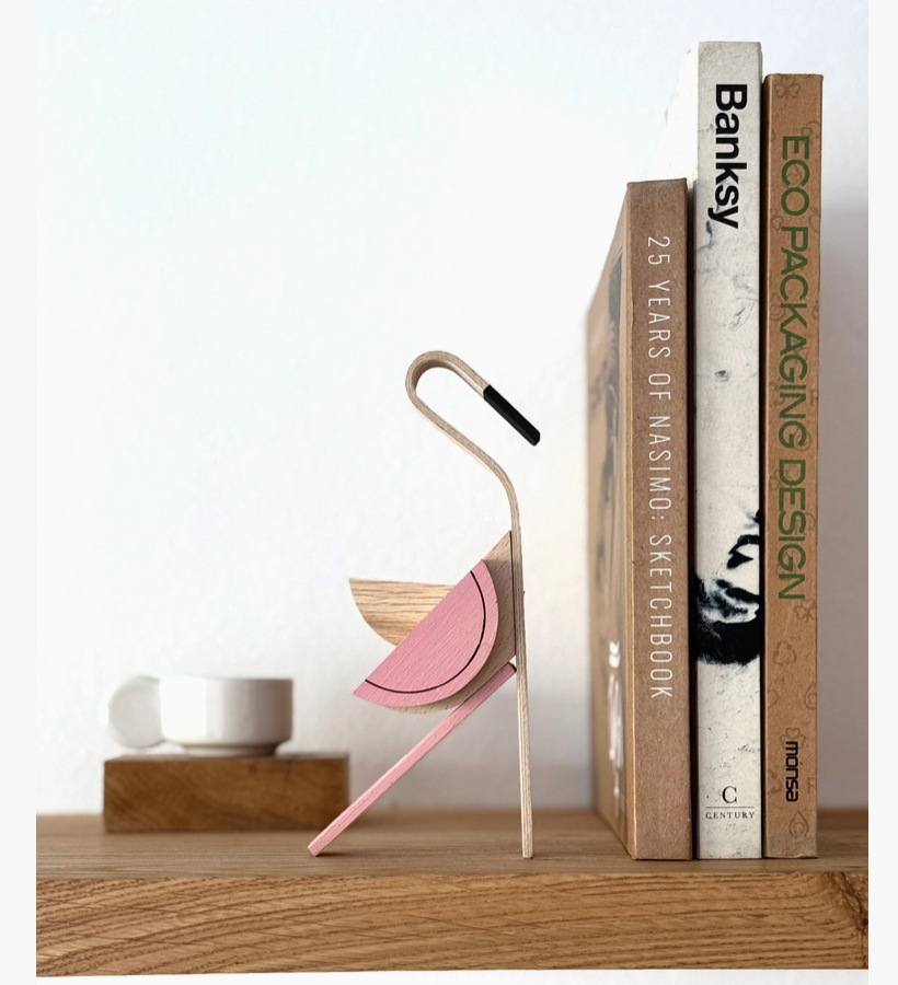 A modern, minimalist wooden flamingo on a wooden shelf next to three books. The bookend features a light natural wood body with a pink curved wing and a black curved beak.