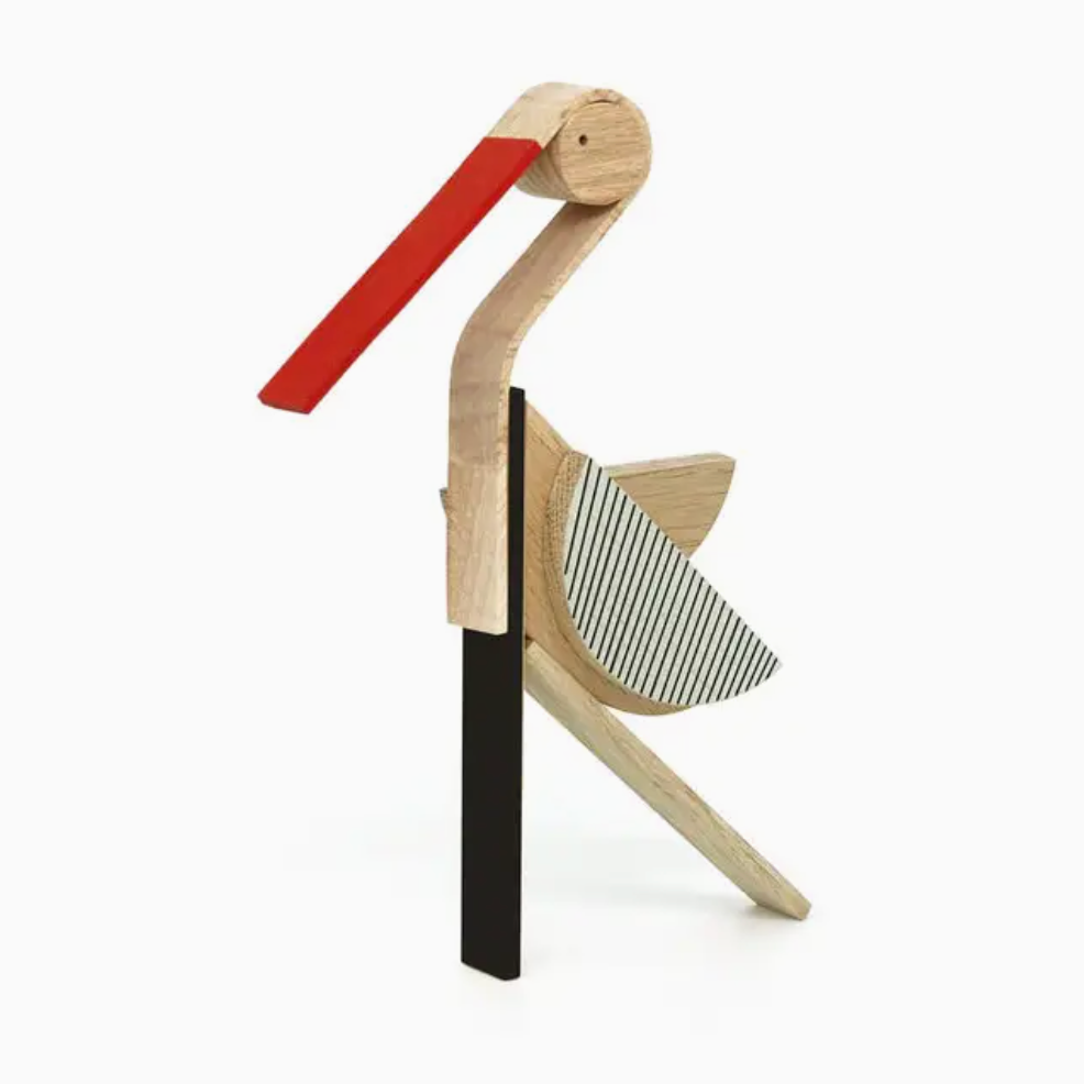 A minimalist wooden bird sculpture or decorative object with a light natural wood body, a long red beak, a black vertical support, and a striped gray-and-white wing, shown on a clean white background.