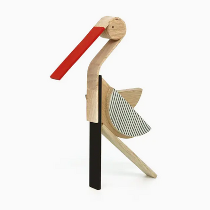 A minimalist wooden bird sculpture or decorative object with a light natural wood body, a long red beak, a black vertical support, and a striped gray-and-white wing, shown on a clean white background.