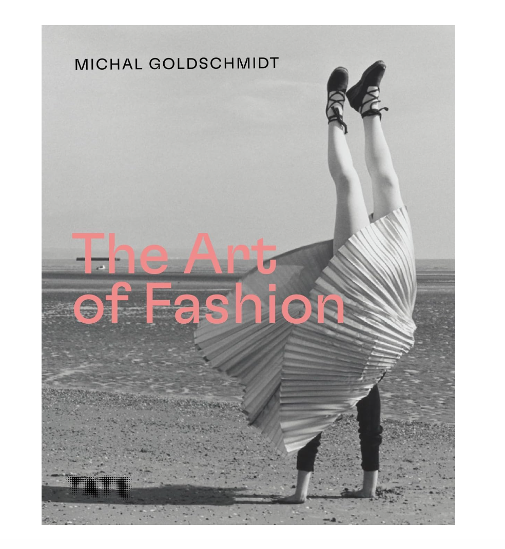 Book cover for 'The Art of Fashion' by Michal Goldschmidt, featuring a black and white photograph of a person on a beach with their legs kicked up into the air, wearing a pleated skirt and black shoes. The title is written in large pink text across the center.