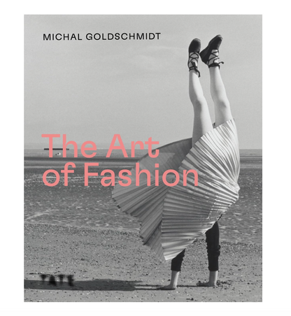 Book cover for 'The Art of Fashion' by Michal Goldschmidt, featuring a black and white photograph of a person on a beach with their legs kicked up into the air, wearing a pleated skirt and black shoes. The title is written in large pink text across the center.