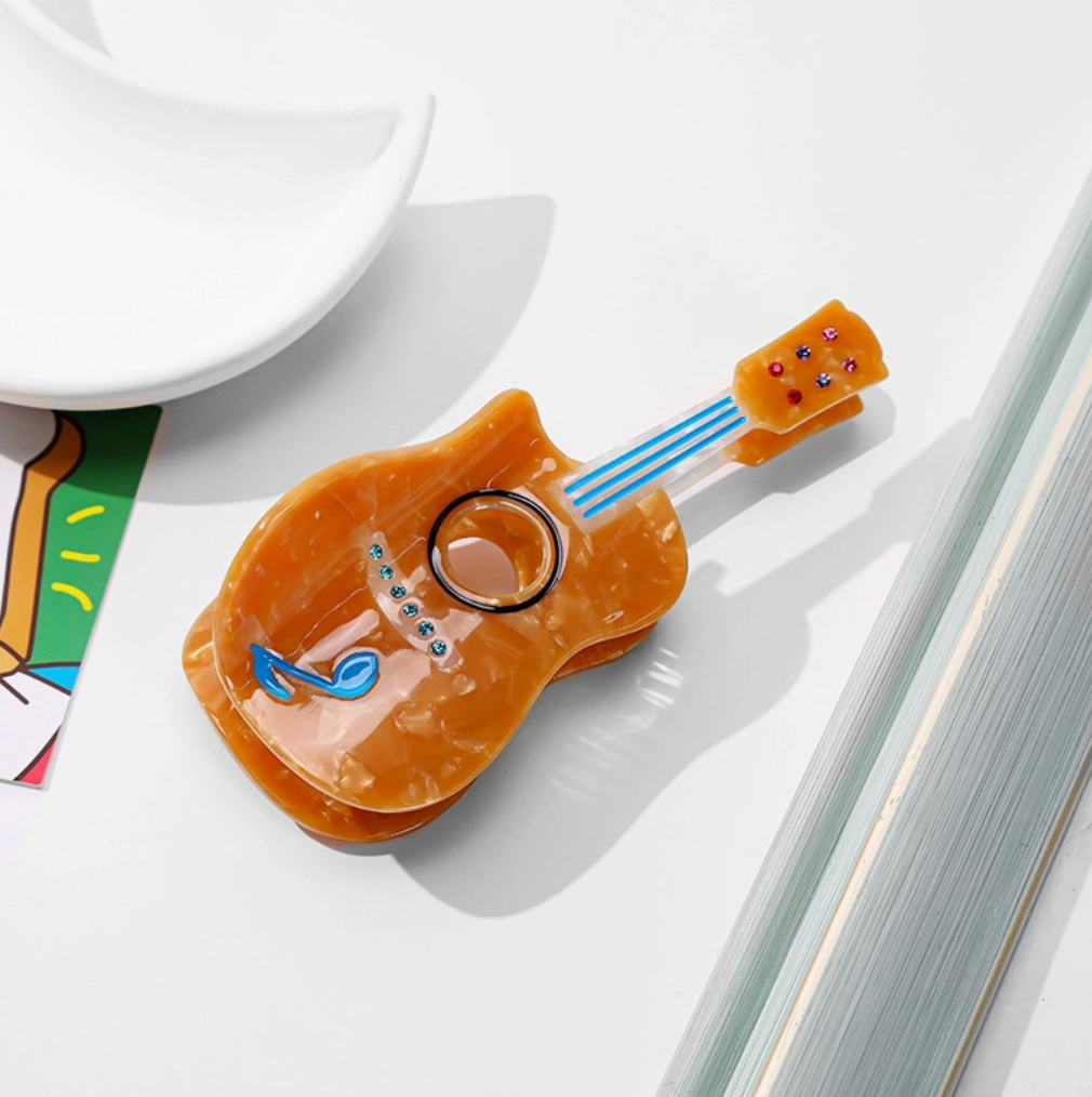 An orange acoustic guitar-shaped hair clip with blue laces and decorative details, displayed on a clean white surface next to a white dish and colorful card.