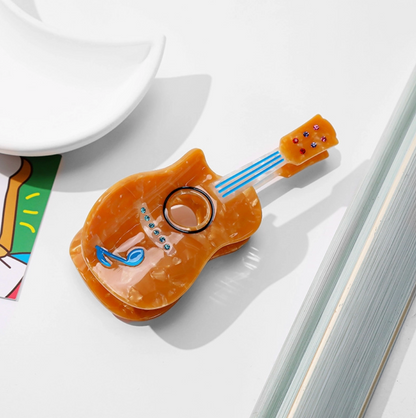 An orange acoustic guitar-shaped hair clip with blue laces and decorative details, displayed on a clean white surface next to a white dish and colorful card.