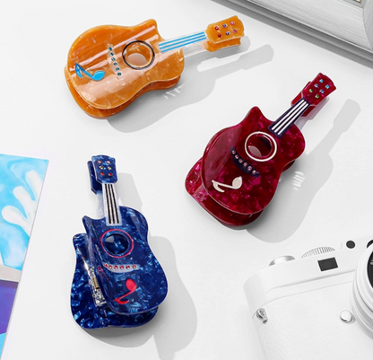 Three miniature guitar-shaped hair clips in translucent orange, red, and blue marbled finishes arranged on a white surface with a camera and paper props.