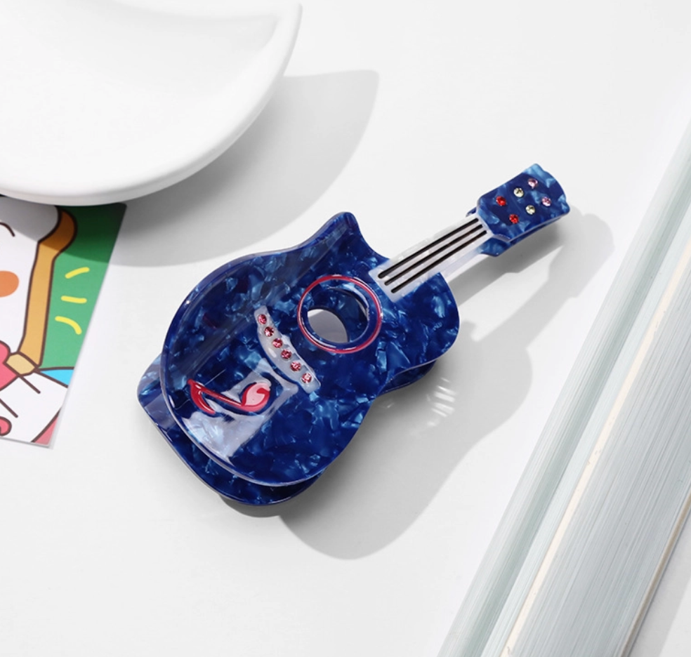 A small guitar-shaped hair clip with a marbled blue and purple body, black neck, and colorful tuning pegs, photographed on a clean white surface with a white plate and colorful card in the background.