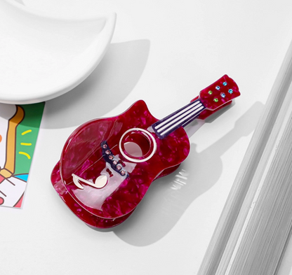 A glossy red guitar-shaped hair clip with a marbled pattern, featuring a circular cutout in the center and small colorful embellishments on the neck, photographed on a white surface with partial props around it.