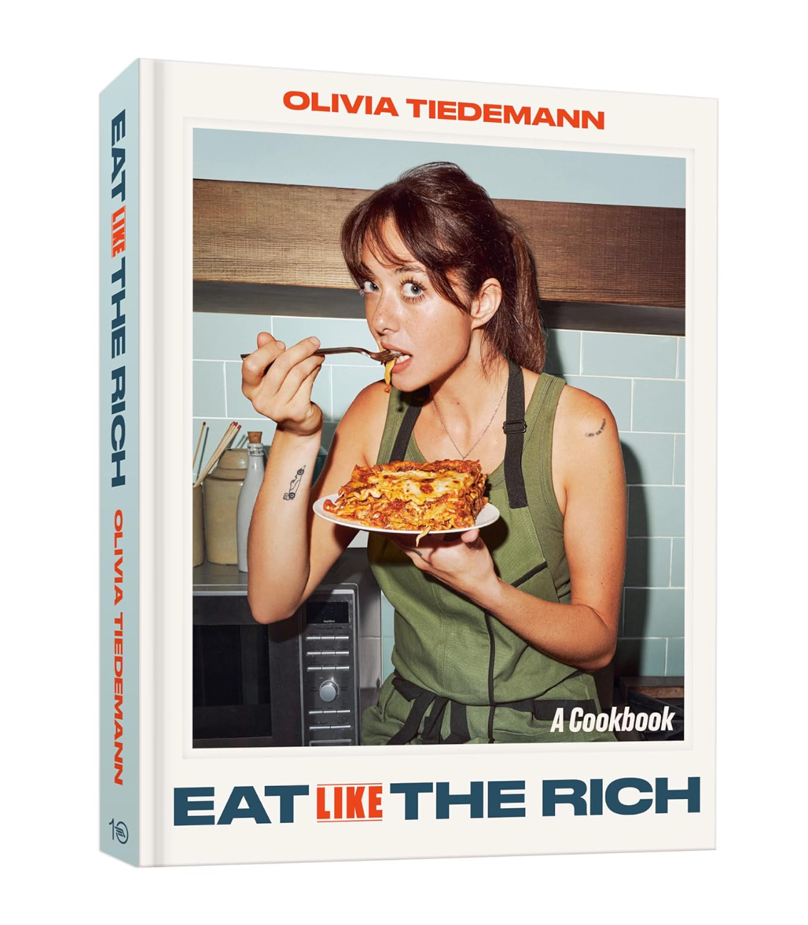 The front and spine of the cookbook 'Eat Like The Rich' by Olivia Tiedemann, featuring a photo of a woman eating pizza in a kitchen on the cover.