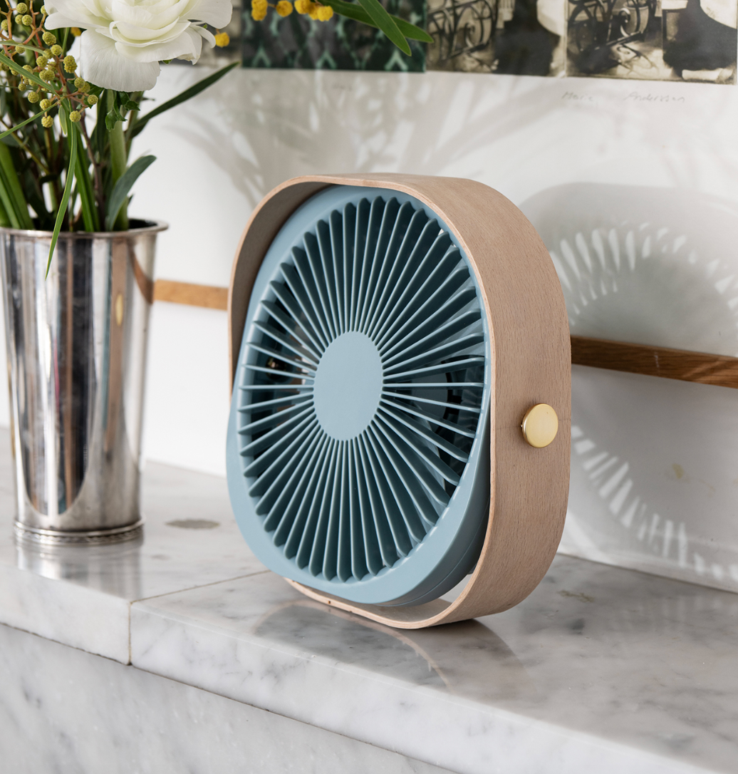 A modern portable fan with a light blue face and a natural wood-grain side frame, resting on a marble countertop next to a silver vase of flowers.