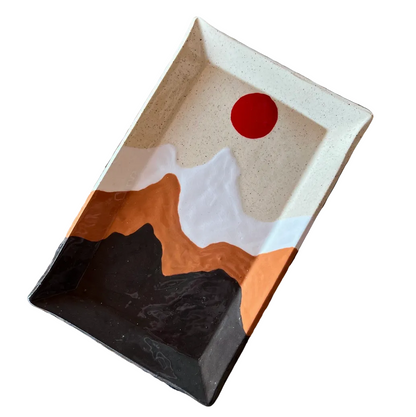 A rectangular ceramic tray with a geometric mountain design in black, white, terracotta orange, and beige, featuring a red sun motif inside, displayed on a woven straw mat.